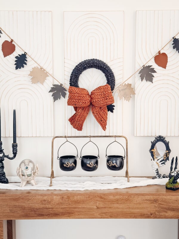 A mantel decorated with a large black Wheat Stitch Crochet Wreath Pattern featuring an orange bow, a garland of paper leaves, three small black cauldrons, and various Halloween-themed figurines.