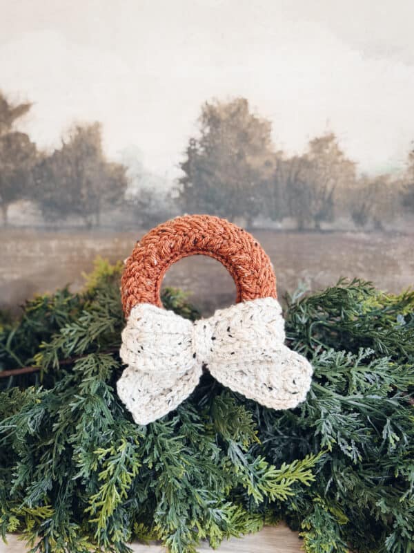 A small Wheat Stitch Crochet Wreath Pattern with a white bow sits on green foliage, with a blurred landscape painting in the background.