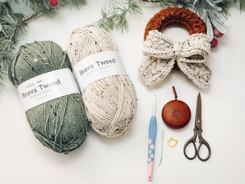 Two skeins of yarn, a crochet hook, tapestry needle, stitch marker, scissors, tape measure, and a crocheted bow wreath are arranged on a white surface with pine branches.