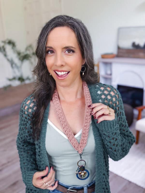 A woman with long brown and gray hair smiles at the camera, wearing a green cardigan, light green top, and a pink lanyard with a bell pendant in a living room setting.