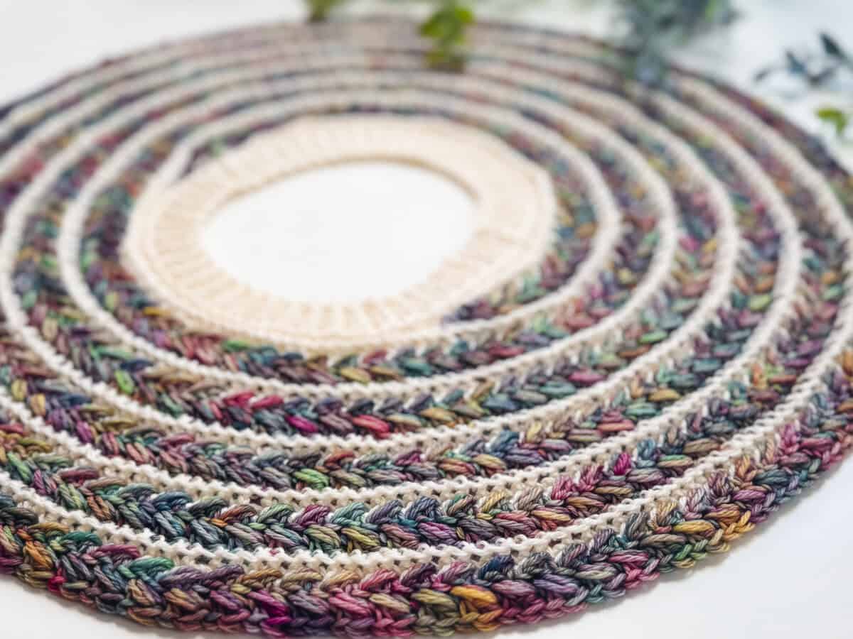 A close-up of a circular, multicolored crocheted mat with concentric rings of variegated and cream yarn, reminiscent of patterns found in a Wheat Stitch Crochet Sweater, laid flat on a white surface.