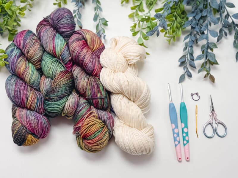 Four skeins of yarn in multicolor and cream, crochet hooks, a needle, stitch marker, and small scissors on a white surface with green foliage at the top.