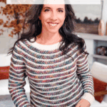 Woman wearing a multicolored wheat stitch crochet sweater stands indoors, smiling. Text highlights a crochet pattern and video tutorial for the sweater, available in sizes XS to 5X.