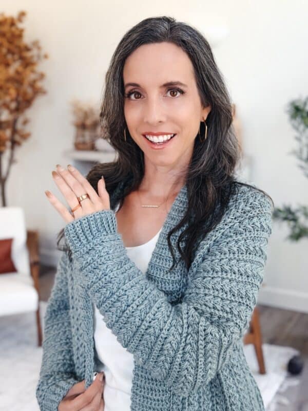 Woman with long brown hair wearing a blue Wheat Stitch Crochet Sweater and gold hoop earrings, smiling and showing rings on her hand, standing in a bright room with plants and decor.