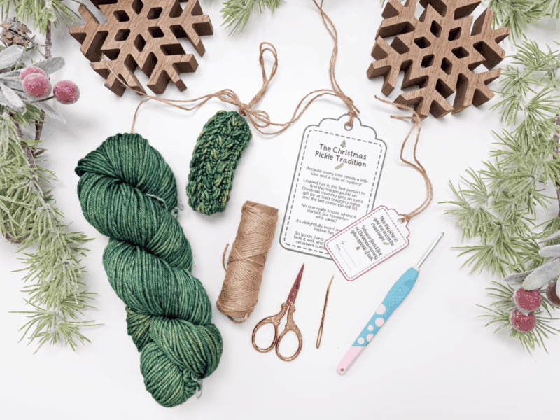 Green yarn, twine, scissors, a crochet hook, a yarn needle, Christmas ornaments, greenery, and a tag explaining the Christmas pickle tradition are arranged on a white surface.