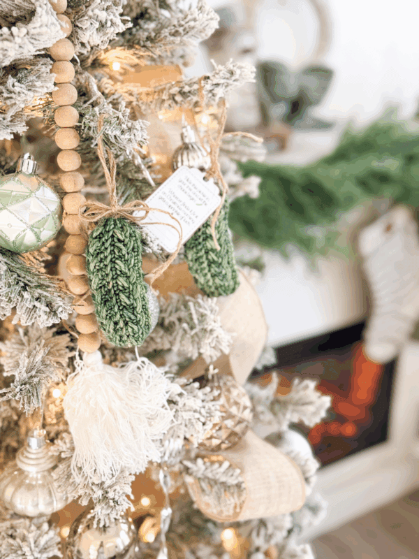 A decorated Christmas tree with green knit mitten ornaments, wooden bead garland, and lights, with a fireplace and greenery visible in the background.