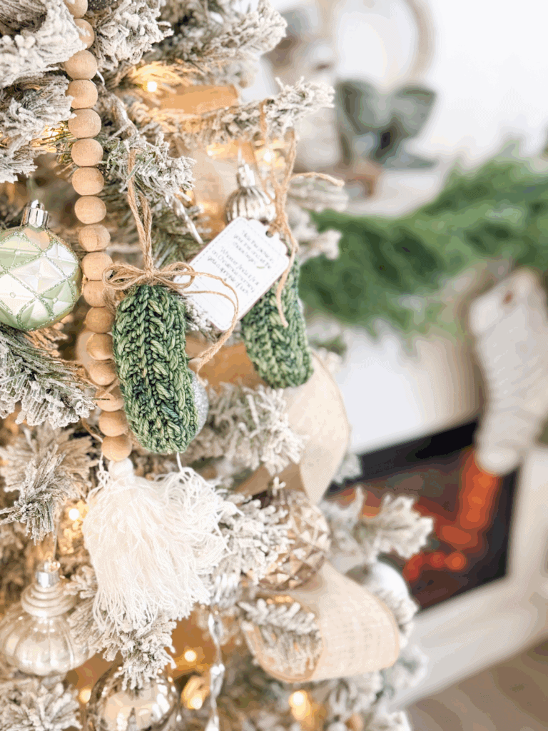 A decorated Christmas tree with green knit mitten ornaments, wooden bead garland, and lights, with a fireplace and greenery visible in the background.