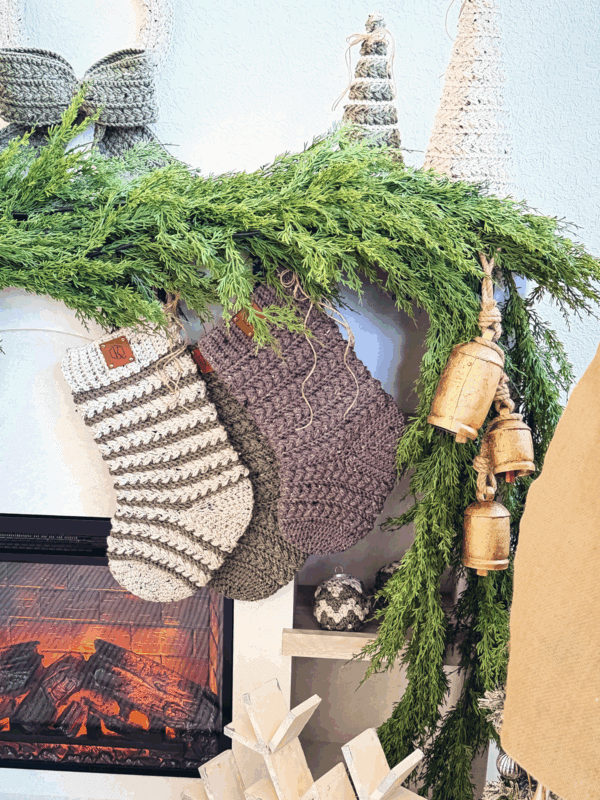 Three knit Christmas stockings hang from a mantel decorated with evergreen garland and golden bells above an electric fireplace with a visible flame effect.