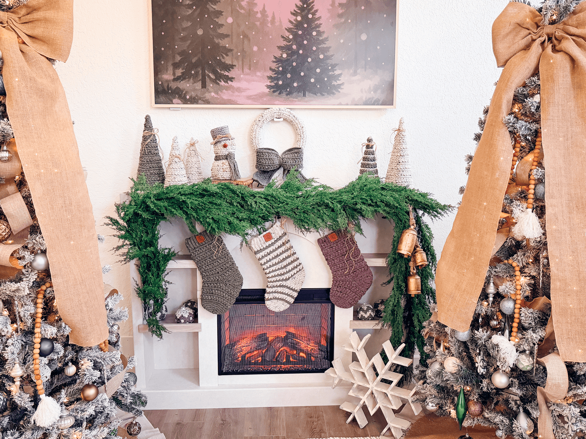 A decorated fireplace mantel with green garland, Wheat Stitch Crochet stockings, small trees, and a wreath, flanked by two Christmas trees with large burlap bows.