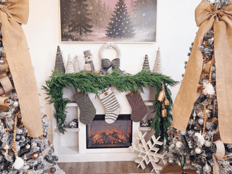 A decorated fireplace with green garland, Wheat Stitch Crochet Stockings, and ornaments, flanked by two Christmas trees with burlap bows and gold decorations. A winter painting hangs above the mantel.