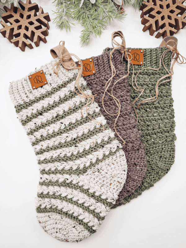 Three crocheted Christmas Wheat Stitch stockings in cream, green, and brown tones with leather tags and twine bows, arranged on a white surface with wooden snowflake decorations and greenery.