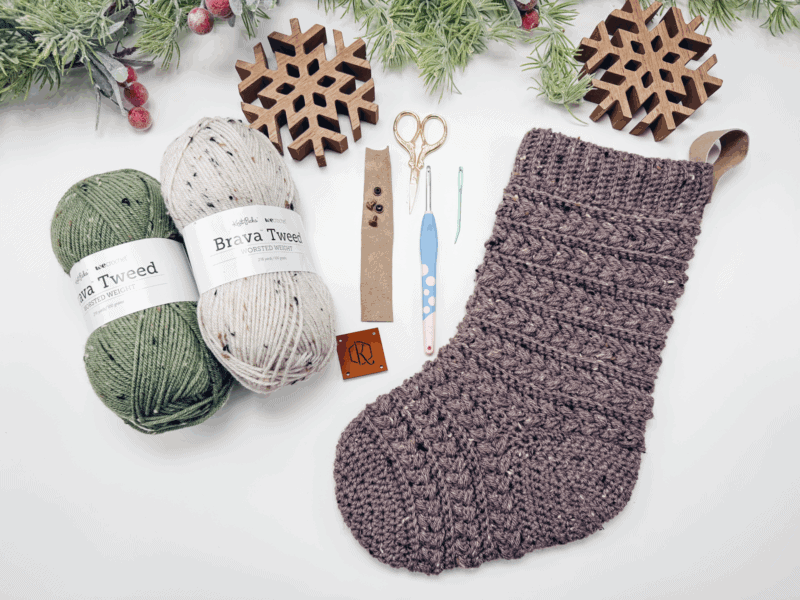 Flat lay of green and beige yarn, brown crocheted Christmas stocking, wooden snowflakes, crochet hooks, scissors, needles, buttons, and pine branches with red berries.