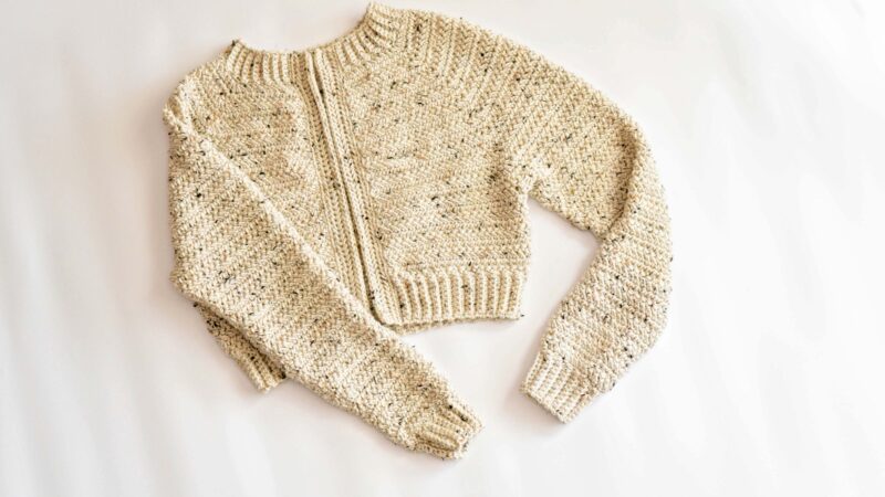 A beige, hand-knitted, long-sleeve cropped sweater inspired by the Cascade Adult Crochet Cardigan design, with a ribbed hem and cuffs, laid flat on a white surface.