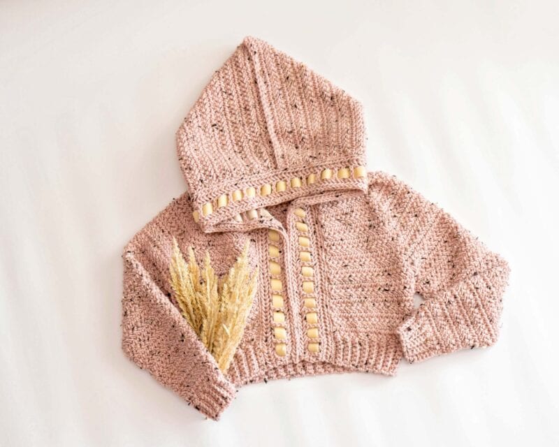 A knitted pink hooded baby sweater with cream-colored buttons and decorative wheat stalks placed on the left side, displayed on a white background&mdash;an adorable complement to a Cascade Adult Crochet Cardigan for matching family style.
