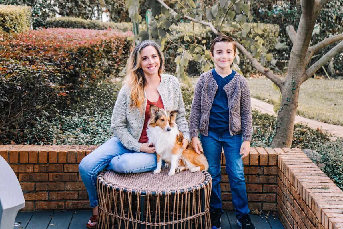 A woman and a boy, with the boy wearing a Cascade Child Crochet Cardigan pattern, stand by an outdoor wicker table where a small dog sits, all surrounded by greenery and brick edging.
