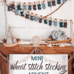 A wooden table decorated with yarn trees and garland sits below three wall art pieces and two hanging rows of Wheat Stitch Crochet Mini Stocking advent displays.