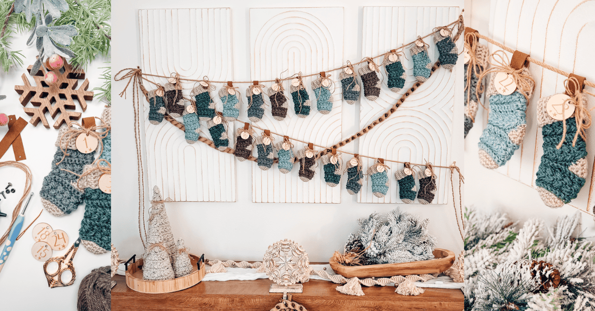 A handmade advent calendar with small crocheted stockings in blue and gray tones hangs on a wall above a wooden table with holiday decorations and greenery.