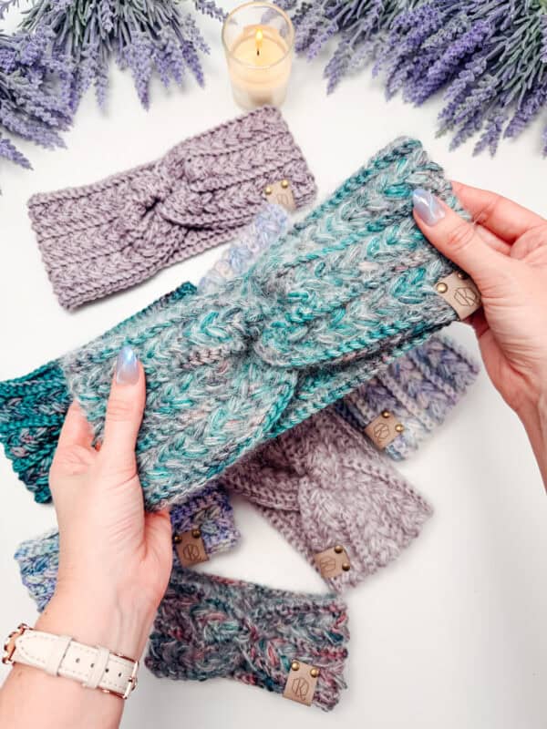 A person holds a knitted green and gray headband above a table displaying several other handmade knit headbands, with a lit candle and purple flowers in the background.