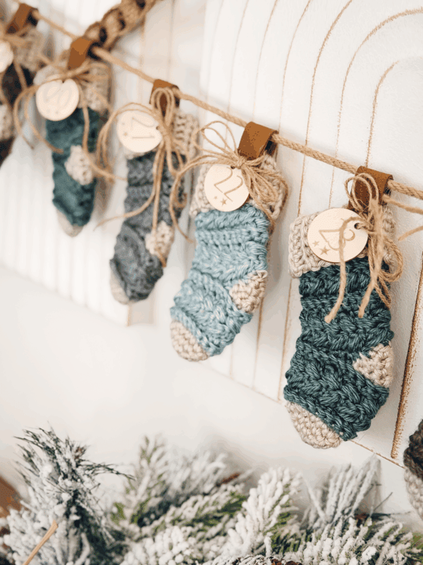 Several small, knitted Christmas stockings hang from a string with numbered wooden tags above frosted greenery.