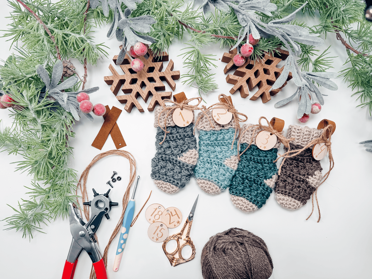 Four small crocheted stockings with numbered wooden tags are arranged under frosted greenery, with crafting tools and yarn on a white surface.