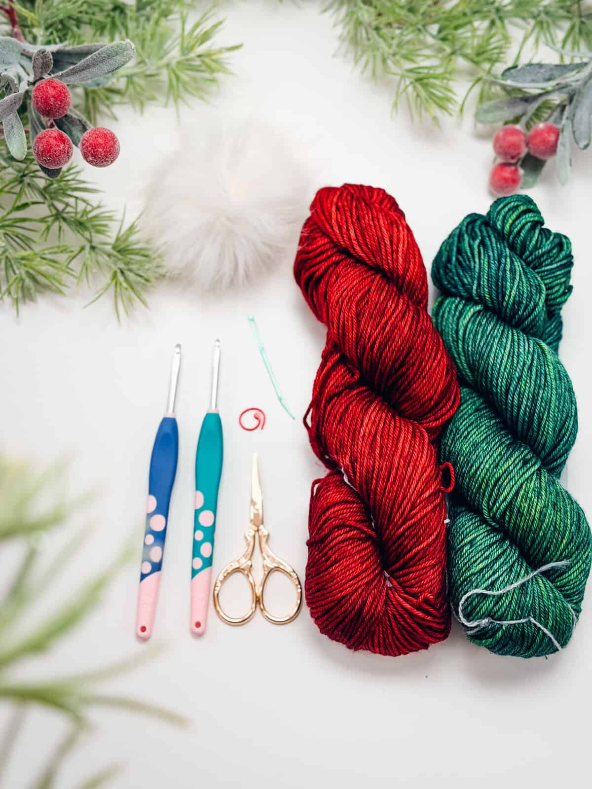 Two skeins of yarn, one red and one green, crochet hooks, scissors, a pom-pom, and stitch markers arranged on a white surface with festive greenery.