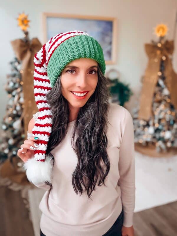 A woman with long dark hair wears a green and red striped holiday hat and a light sweater, standing in front of decorated Christmas trees.