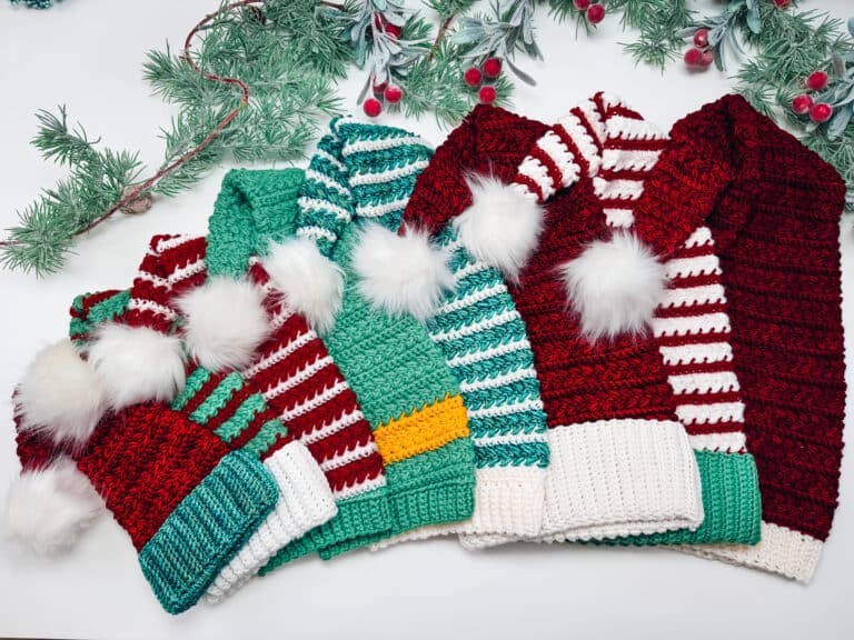 Six crocheted winter hats with stripes and pom-poms are arranged in a row on a white surface, with pine branches and red berries above them.