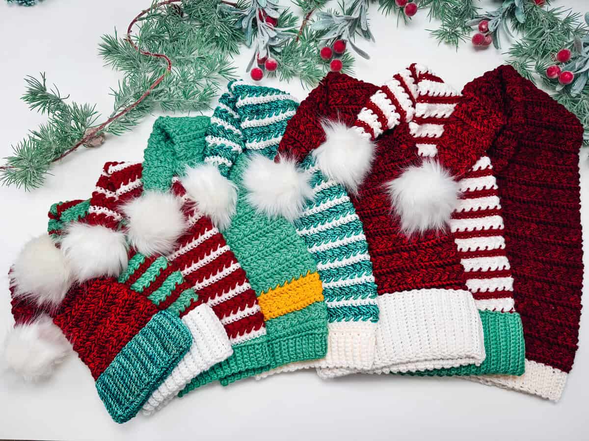 Six crocheted winter hats with stripes and pom-poms are arranged in a row on a white surface, with pine branches and red berries above them.