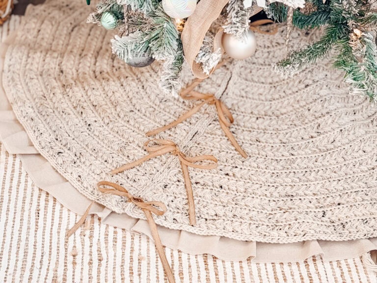 A close-up of a braided tree skirt with ribbon bows under a decorated Christmas tree, on a textured striped rug.