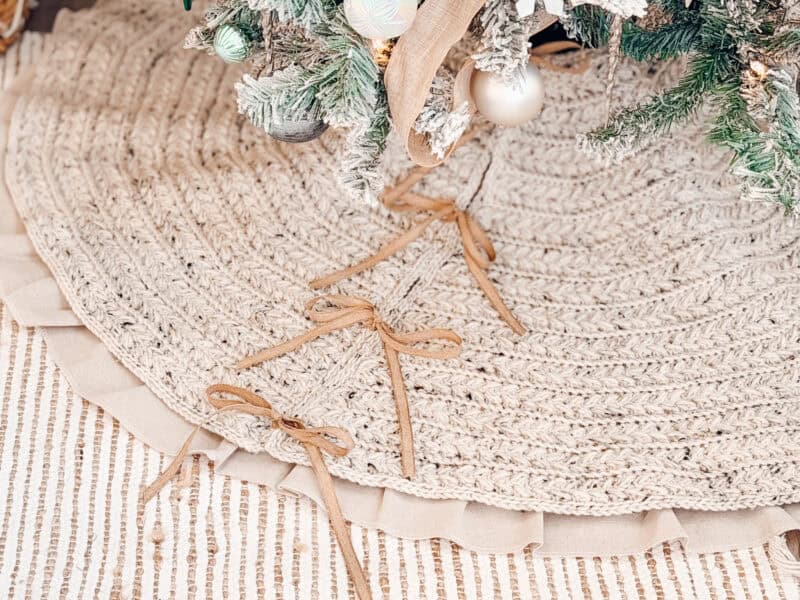 A close-up of a braided tree skirt with ribbon bows under a decorated Christmas tree, on a textured striped rug.