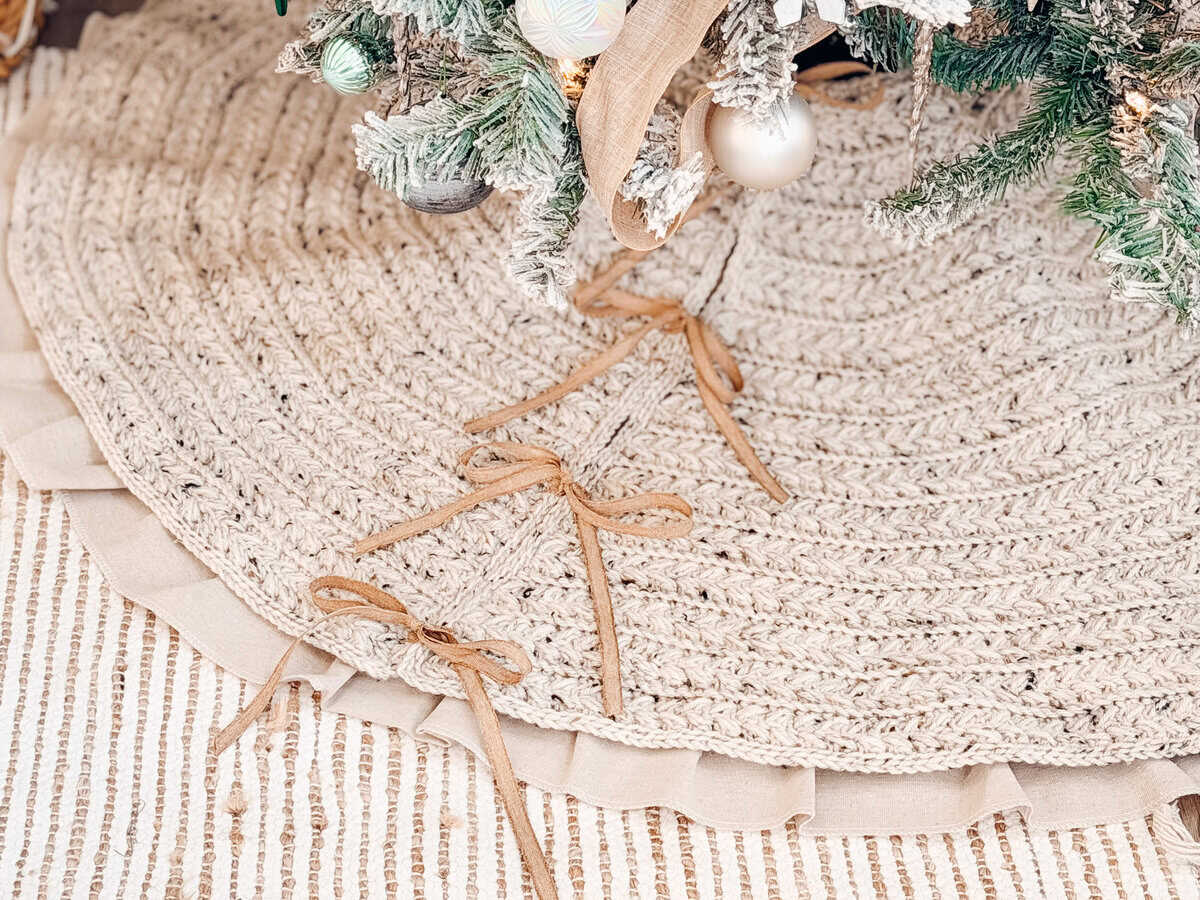 A close-up of a braided tree skirt with ribbon bows under a decorated Christmas tree, on a textured striped rug.