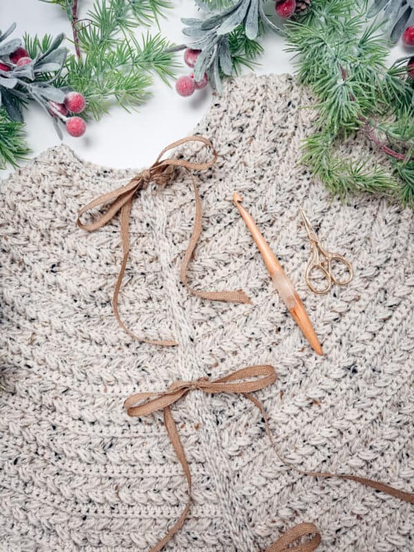 Close-up of a beige crocheted garment using the Wheat Stitch Crochet Tree Skirt Pattern with brown ties, a wooden crochet hook, and gold scissors, surrounded by artificial evergreen branches and red berries.
