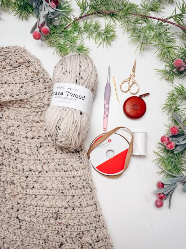 Flat lay of crochet supplies including tweed yarn, a crochet hook, scissors, measuring tape, thread, and a partially completed crochet project, with decorative greenery and berries.