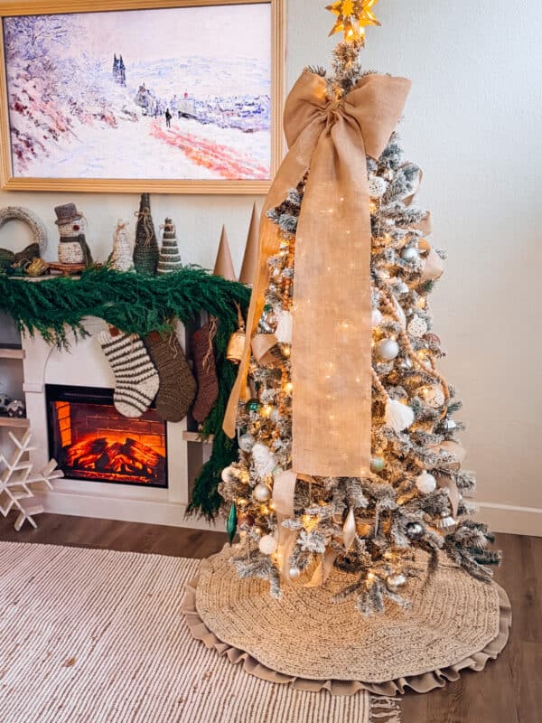 A decorated Christmas tree with a large burlap bow stands next to a fireplace adorned with stockings, greenery, and a holiday painting above the mantel, finished with a Wheat Stitch Crochet Tree Skirt Pattern for a cozy touch.