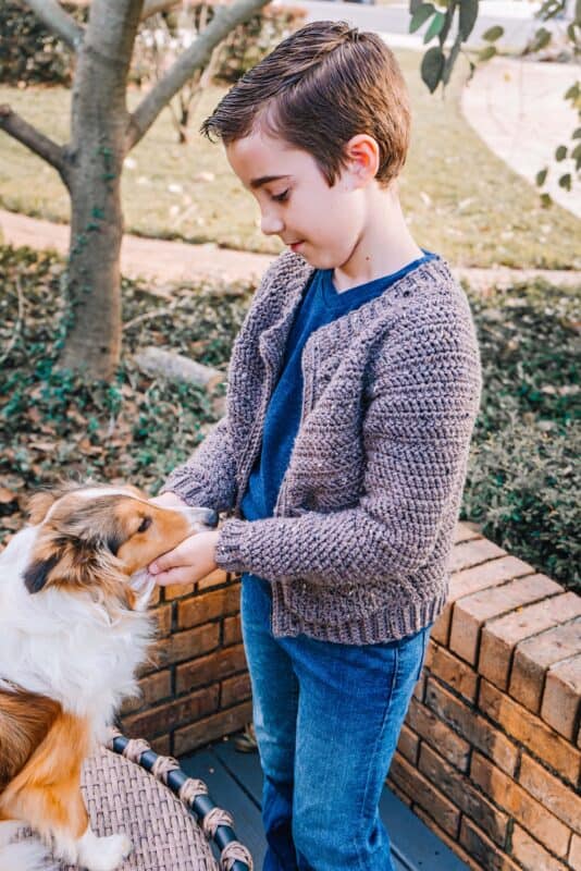 A young boy in a Cascade Adult Crochet Cardigan and jeans gently pets a small dog outdoors near a brick wall and green bushes.