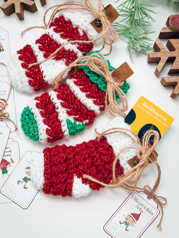 Three Wheat Stitch Crochet Mini Stockings in red, white, and green, each tied with twine&mdash;one holding a gift card. Wooden decorations and gift tags are arranged nearby for a festive touch.