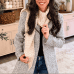 Woman wearing a gray coat and cream hooded crochet scarf, standing indoors by shelves with yarn and decor. Text reads “Wheat Stitch™ Crochet Hooded Scarf Pattern www.brianakdesigns.com”.