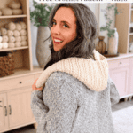 Woman wearing a gray coat with a cream-colored crocheted hooded scarf, standing in a cozy room with shelves of yarn and decor. Text reads: “Wheat Stitch™ crochet hooded scarf pattern – Free Crochet Pattern + Video.”.