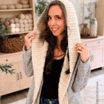 Woman wearing a crocheted Wheat Stitch™ crochet hooded scarf pattern, standing indoors, holding up the hood; yarn and plants are displayed on shelves in the background.