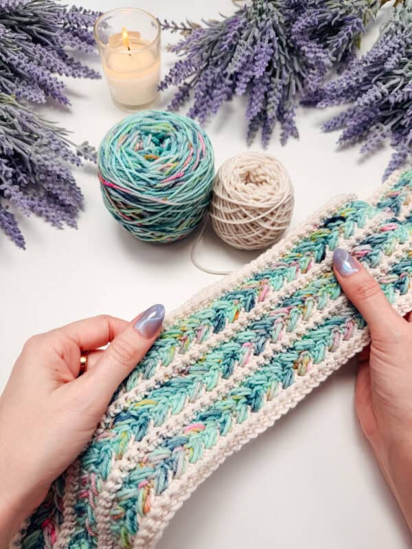 Hands holding a crocheted piece with pastel blue and white yarn, surrounded by yarn balls, lavender sprigs, and a lit candle on a white surface.