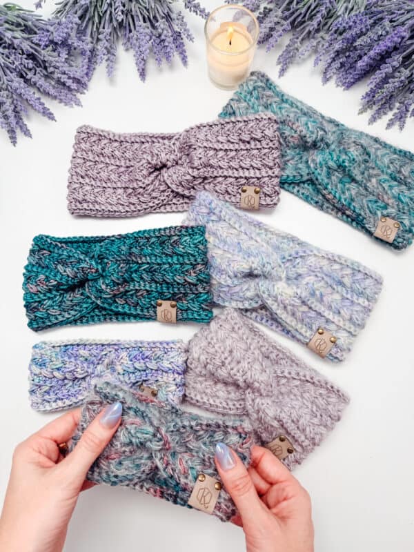 Six knitted headbands in various shades of purple, blue, and teal are displayed on a white surface with lavender, a candle, and two hands holding one headband.