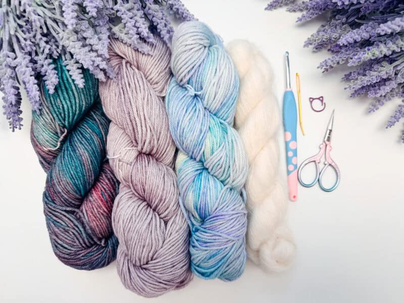 Four skeins of multicolored yarn, crochet hooks, scissors, and a needle arranged on a white surface with lavender flowers in the corners.