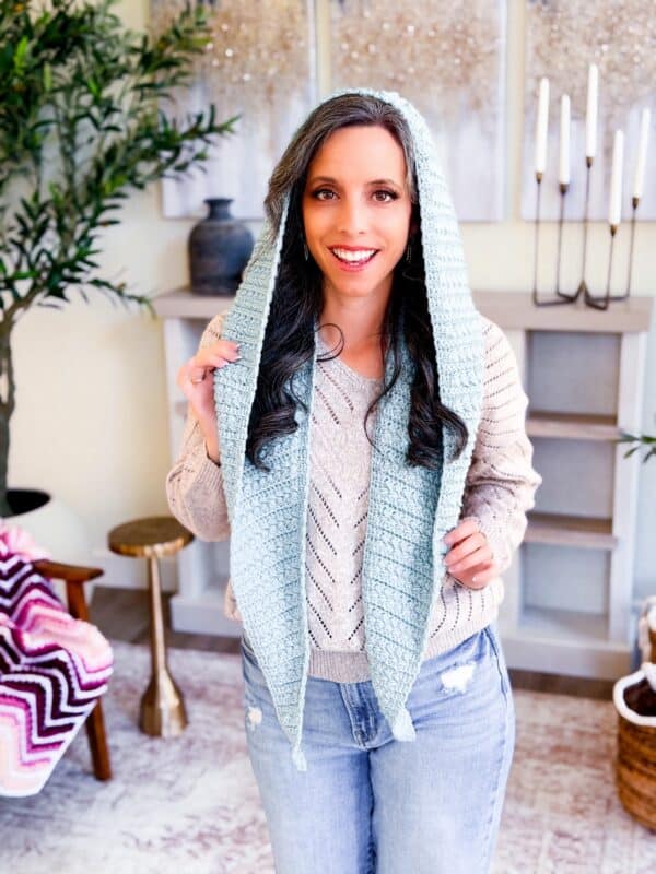 A woman wearing a light blue Wheat Stitch&trade; crochet hooded scarf stands indoors, smiling at the camera. She pairs it with a beige sweater and jeans, while stylish home decor accents the background.