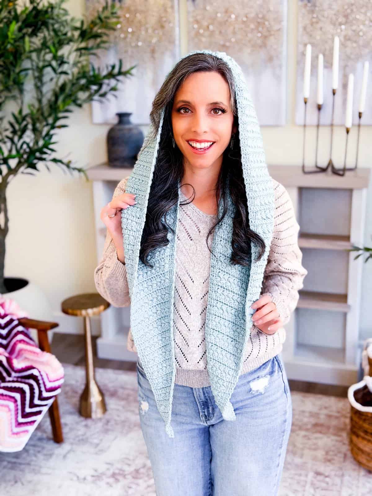 A woman wearing a light blue Wheat Stitch™ crochet hooded scarf stands indoors, smiling at the camera. She pairs it with a beige sweater and jeans, while stylish home decor accents the background.