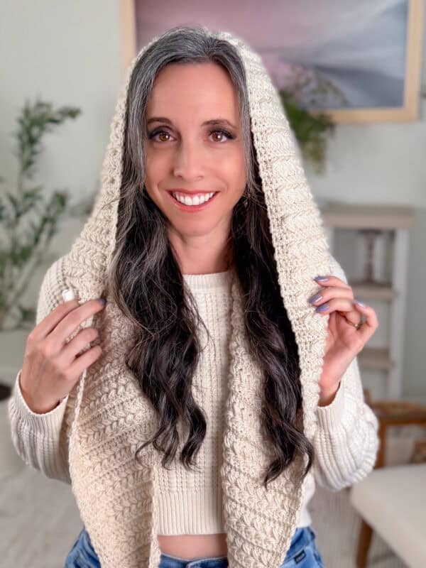 A woman with long, wavy dark hair and gray highlights smiles while holding a knitted cream-colored scarf draped over her head. She wears a cream sweater and blue jeans.