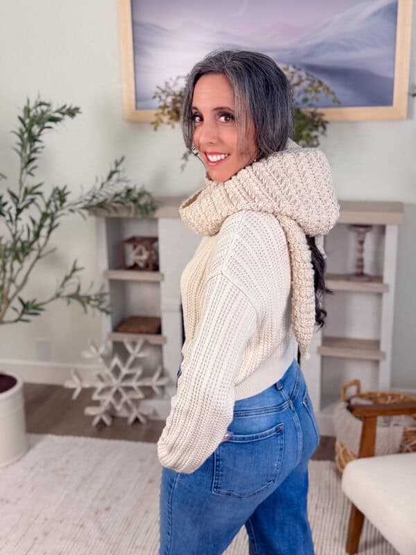 A woman with long gray hair wearing a cream knit sweater and scarf stands indoors, smiling, with her head turned over her shoulder.