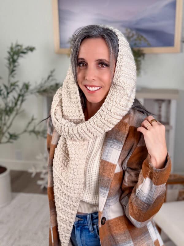 A woman wearing a plaid coat, cream knit scarf and hood, and a cream sweater stands indoors, smiling at the camera.