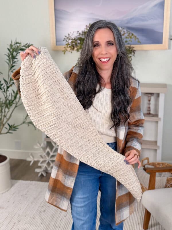 A woman with long gray hair, wearing a plaid coat and jeans, holds up a textured, off-white crocheted piece in a cozy, well-decorated room.