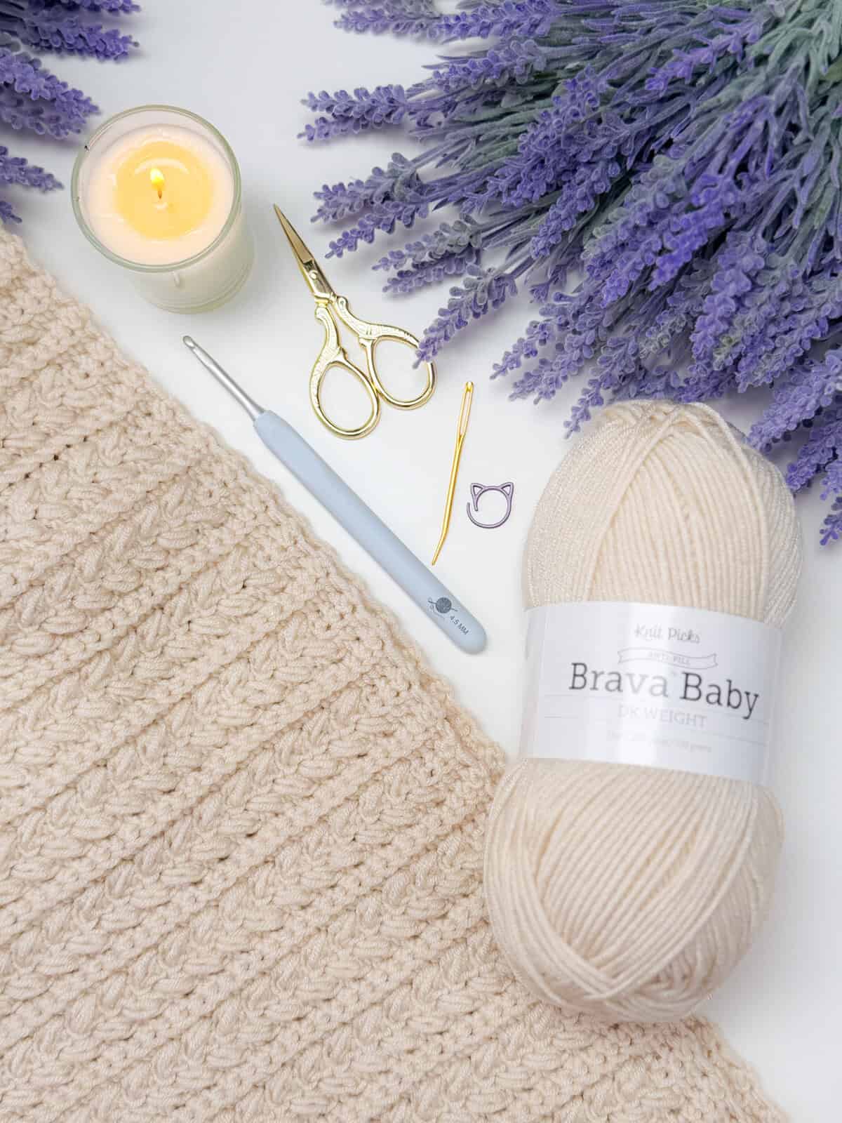 A cream-colored crochet blanket, a skein of Brava Baby yarn, crochet hook, gold scissors, yarn needle, stitch marker, candle, and lavender sprigs arranged on a white surface.