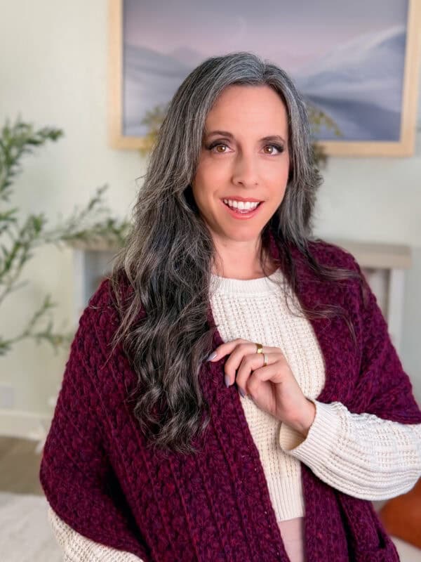 Woman with long gray hair wearing a cream sweater and maroon shawl, smiling and standing indoors with a landscape painting in the background.